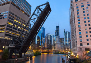 An image of the Kinzie Street Rail Bridge over the Chicago River, with the Willis Tower in the background. The bridge is in an upright position over the Chicago River to allow boats to pass.
